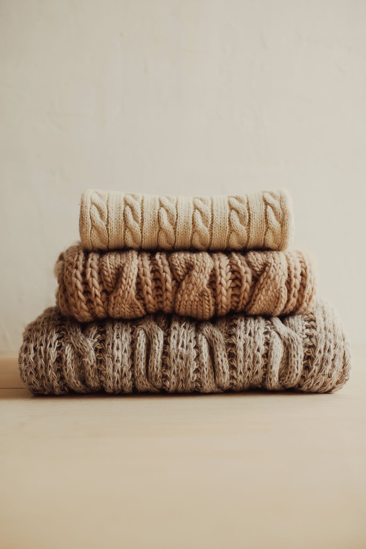 Three folded chunky knit pullovers in oat, camel, and stone, from the illustrative product example